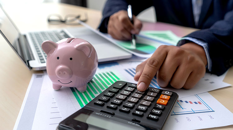 Man uses calculator and laptop to estimate custom software development cost. A piggy bank is next to the laptop.